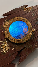 Sculpture made of wood of a ripped part of a ship, left only with a circular window and many barnacles on the side of the wood. Inside the window are floating plastic bags, small jellyfish and coral, behind cracked glass. Close up and backlit with blue lighting.