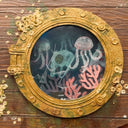 Sculpture made of wood of a ripped part of a ship, left only with a circular window and many barnacles on the side of the wood. Inside the window are floating plastic bags, small jellyfish and coral, behind cracked glass. Close up.