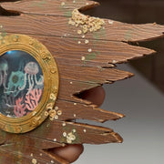 Sculpture made of wood of a ripped part of a ship, left only with a circular window and many barnacles on the side of the wood. Inside the window are floating plastic bags, small jellyfish and coral, behind cracked glass. Close up.