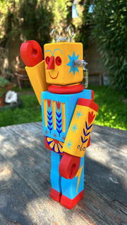 Wooden sculpture of a toy robot, painted in bright yellow, red and blue with floral and starburst patterns. It has a metal coil on its head and lightbulbs coming out the side of its head.