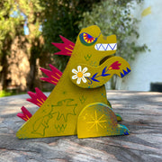 Wooden sculpture of a yellowish green Godzilla, stylized in a folklore styl with floral and starburst detailing. It has burgundy spikes and grins.