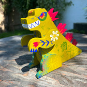 Wooden sculpture of a yellowish green Godzilla, stylized in a folklore styl with floral and starburst detailing. It has burgundy spikes and grins.