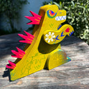 Wooden sculpture of a yellowish green Godzilla, stylized in a folklore styl with floral and starburst detailing. It has burgundy spikes and grins.