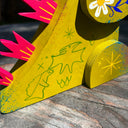 Wooden sculpture of a yellowish green Godzilla, stylized in a folklore styl with floral and starburst detailing. It has burgundy spikes and grins.