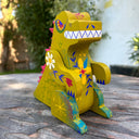 Wooden sculpture of a yellowish green Godzilla, stylized in a folklore styl with floral and starburst detailing. It has burgundy spikes and grins.