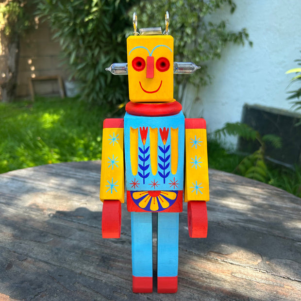Wooden sculpture of a toy robot, painted in bright yellow, red and blue with floral and starburst patterns. It has a metal coil on its head and lightbulbs coming out the side of its head.
