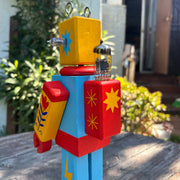 Wooden sculpture of a toy robot, painted in bright yellow, red and blue with floral and starburst patterns. It has a metal coil on its head and lightbulbs coming out the side of its head.