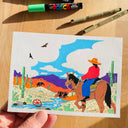 Artwork featuring a horseback rider on a horse in a desert scene, held by a hand on a wooden surface with markers.
