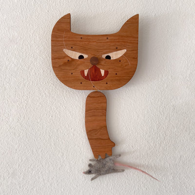 Die cut wooden sculpture, of a cat's head with a swinging wooden cat arm below it. A felt dead mouse is attached to the cats paw and the face of the cat resembles a simplified clock.