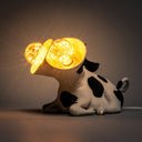 Lamp designed as a cow's body sitting on the ground, with 2 light bulbs coming from 2 separate cow heads conjoined to the same neck.