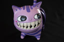 Ceramic sculpture of the Cheshire cat, stylized to have large human like teeth and an empty coil for a body.  It looks off to the side with a slightly mischievous look.