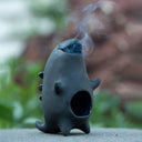 Ceramic kaiju-shaped incense burner with smoke emerging against a blurred natural background