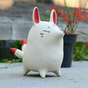 A small white ceramic fox figurine with red inner ears, red cheek marks, and red-tipped tail quills, standing outdoors on a concrete surface with a blurred wooden fence and potted plant in the background.