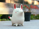 A small white ceramic fox figurine with red inner ears, red cheek marks, and red-tipped tail quills, standing outdoors on a concrete surface with a blurred wooden fence and potted plant in the background.