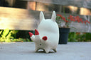 A small white ceramic fox figurine with red inner ears, red cheek marks, and red-tipped tail quills, standing outdoors on a concrete surface with a blurred wooden fence and potted plant in the background.