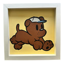 A square framed painting featuring a cartoon brown puppy wearing a white and blue cap, set against a yellow background and enclosed in a white frame.