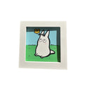 Framed illustration of a very small white chibi Totoro character, sitting on grass with a yellow flower atop its head.