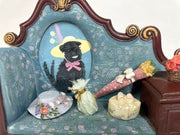 Painting of a small black dog in a wide brimmed yellow hat with a pink feather coming out of it. Its framed in a 3D sculptural frame of a couch with little knick knacks on it.