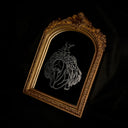 An ornate gold arched frame containing a mirror with a white line-art engraving of an angel hunched over, with her head dropped down in dismay and viscera exploding out her back, photographed against a black backdrop.
