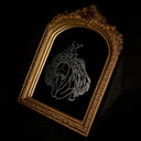 An ornate gold arched frame containing a mirror with a white line-art engraving of an angel hunched over, with her head dropped down in dismay and viscera exploding out her back, photographed against a black backdrop.