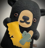 Flat plush black bear, with shiny eyes and short limbs. It holds up a plush cut out of California and a small cactus.