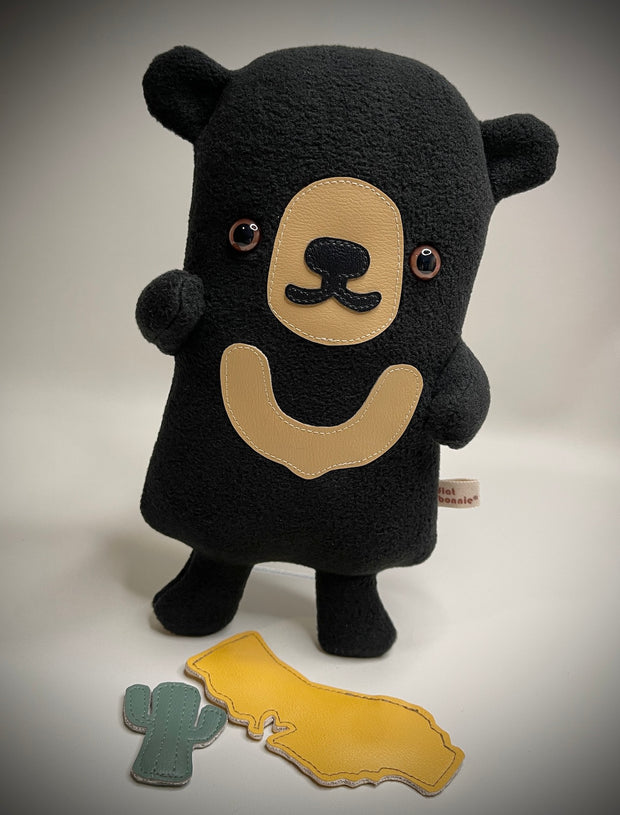 Flat plush black bear, with shiny eyes and short limbs. It holds up a plush cut out of California and a small cactus.