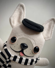 Cream colored plush dog mostly flat with large pointed ears and a sewn on face. It wears a hat and a striped scarf and has its small tongue out, smiling.