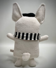 Cream colored plush dog mostly flat with large pointed ears and a sewn on face. It wears a hat and a striped scarf and has its small tongue out, smiling.