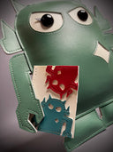 Sage green vinyl fabric plush of a Big Boss Robot, slightly shorter and with large eyes. It holds a small white magazine with a red and a blue robot on it.