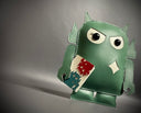 Sage green vinyl fabric plush of a Big Boss Robot, slightly shorter and with large eyes. It holds a small white magazine with a red and a blue robot on it.