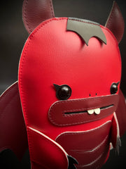 Red vinyl plush of a devil creature with an outlined mouth and buckteeth. It has horns, wings and a fork under one of its arms.
