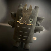 Plush of a black dragon, with a very squarish body and small arms and legs. Its facial features are made of vinyl, with a sharp tooth smile and a striped underbelly.