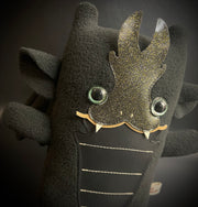 Plush of a black dragon, with a very squarish body and small arms and legs. Its facial features are made of vinyl, with a sharp tooth smile and a striped underbelly.