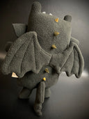 Plush of a black dragon, with a very squarish body and small arms and legs. Its facial features are made of vinyl, with a sharp tooth smile and a striped underbelly. It has gold spikes along its back.