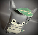 Vinyl plush sculpture of a Totoro inspired character, depicted in a flat style with small round eyes and legs. It has a wide eyed smile and holds a leaf umbrella overhead. 