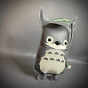 Vinyl plush sculpture of a Totoro inspired character, depicted in a flat style with small round eyes and legs. It has a wide eyed smile and holds a leaf umbrella overhead. 