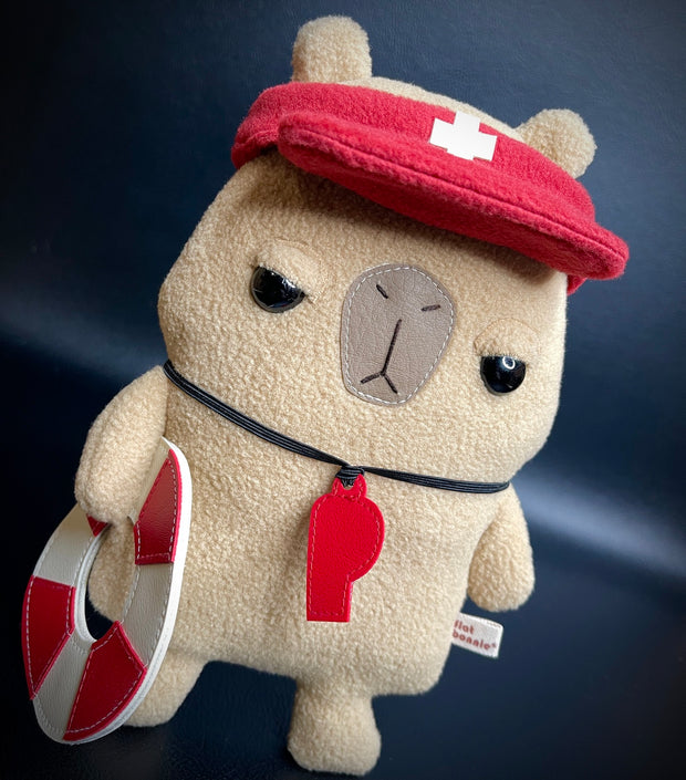 Plush of a capybara with simplistic features and a cartoon facial expression to make it look unimpressed. It holds a life ring, has a red whistle around its neck and a lifeguard visor atop its head.