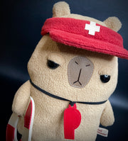 Plush of a capybara with simplistic features and a cartoon facial expression to make it look unimpressed. It holds a life ring, has a red whistle around its neck and a lifeguard visor atop its head.