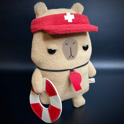 Plush of a capybara with simplistic features and a cartoon facial expression to make it look unimpressed. It holds a life ring, has a red whistle around its neck and a lifeguard visor atop its head. 