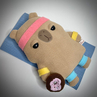 Plush of a flat shaped cartoon capybara, with a frustrated facial expression. It lays on a yoga mat with sweatbands on its head, arms and legs and holds a donut.