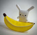 Plush sculpture of a small white bunny sitting on a pleather banana.