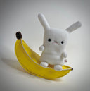 Plush sculpture of a small white bunny sitting on a pleather banana.