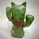 VInyl fabric plush doll of a cactus with a cat face and cat ears. It has metal spikes, a brown fabric bandana and a small pleather pouch.