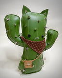 VInyl fabric plush doll of a cactus with a cat face and cat ears. It has metal spikes, a brown fabric bandana and a small pleather pouch.