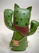 VInyl fabric plush doll of a cactus with a cat face and cat ears. It has metal spikes, a brown fabric bandana and a small pleather pouch.