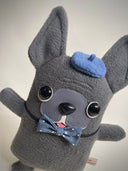 Flat plush doll of a gray French Bull dog with pointy ears, standing on 2 legs like a person. It has black bead eyes and wears a navy bowtie and matching beret cap.