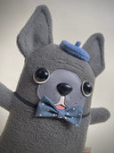 Flat plush doll of a gray French Bull dog with pointy ears, standing on 2 legs like a person. It has black bead eyes and wears a navy bowtie and matching beret cap.