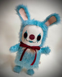 A blue furry bunny costume plush toy with a white face, red nose, black eye makeup, and a red bow tie.