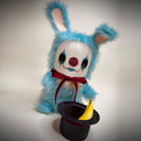 A blue furry bunny costume plush toy with a white face, red nose, black eye makeup, and a red bow tie, posed with a black top hat and a yellow banana in front of a plain studio background.