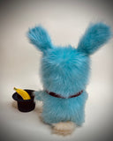 A fluffy light blue bunny plush toy with long fur, standing upright and facing away from the camera. It wears a dark brown collar and is posed next to a small black top hat holding a yellow banana, against a plain light gray studio background.